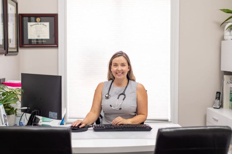Dr. Melanie Hudson, ND, smiling behind her clinic desk with a stethoscope around her neck, ready to welcome a patient.