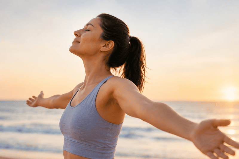 Woman standing by the water with arms open, representing stress relief and emotional well-being