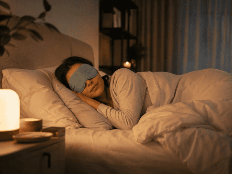 Woman sleeping peacefully while wearing a sleep mask in a dark, calming bedroom.