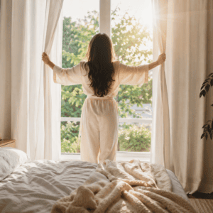 Woman opening bedroom curtains after waking to let in natural morning sunlight.
